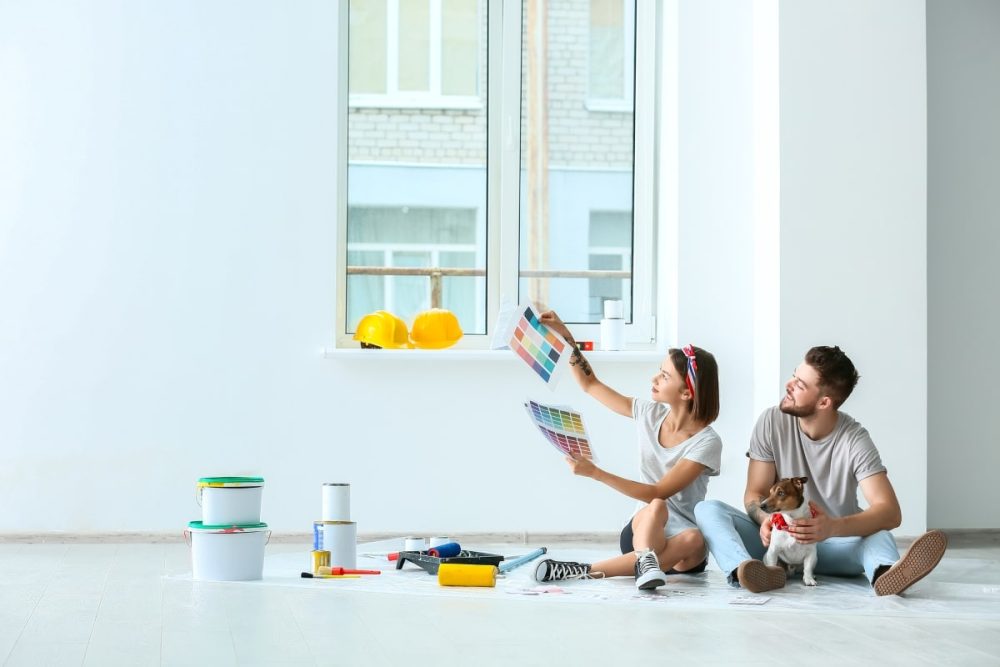 Young couple with their dog, sitting in a room undergoing renovations, choosing paint colors from multiple swatches, indicative of decisions new homeowners face.