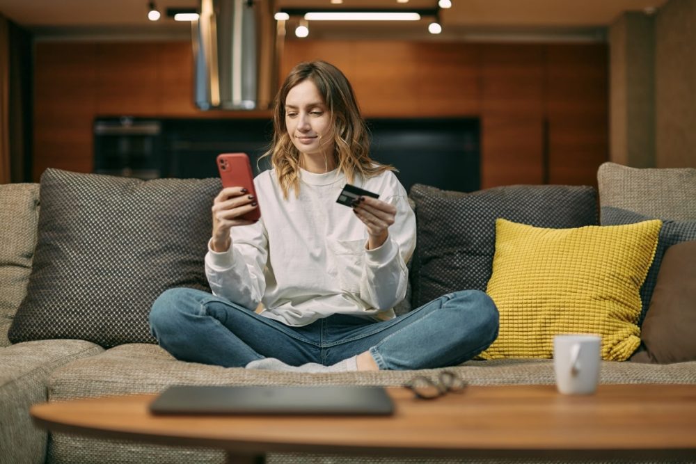A woman sitting comfortably on a couch, paying bills with her phone and credit card, showcasing the financial reliability that landlords look for in a tenant.