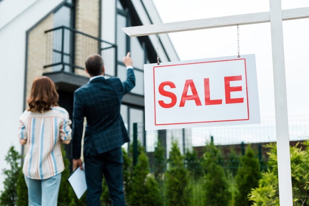 A focused image on a vibrant 'SALE' sign in the foreground, with a potential buyer and a real estate agent blurred in the background pointing towards a modern home. The scene captures the essence of marketing a house for sale, with the staging of the property implicitly suggested by the neat exterior and professional appearance of the agent and client.