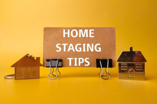 A creative presentation featuring a cardboard sign with the words 'HOME STAGING TIPS' clipped between two small, metallic house-shaped paperclips, all against a vibrant yellow background, symbolising the concept of staging a home for sale.