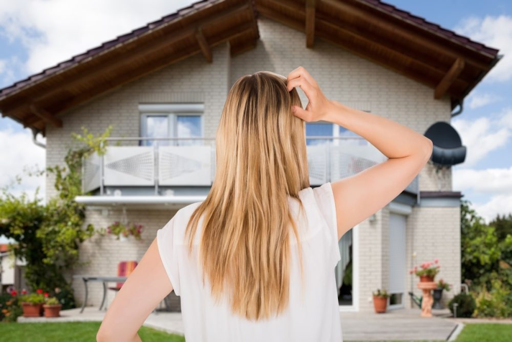 A woman standing with her back to the camera, scratching her head while looking at a two-story house, symbolising the contemplation involved in making a home purchase decision.