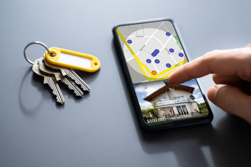 A hand using a real estate search app on a smartphone, with a set of keys on a table.