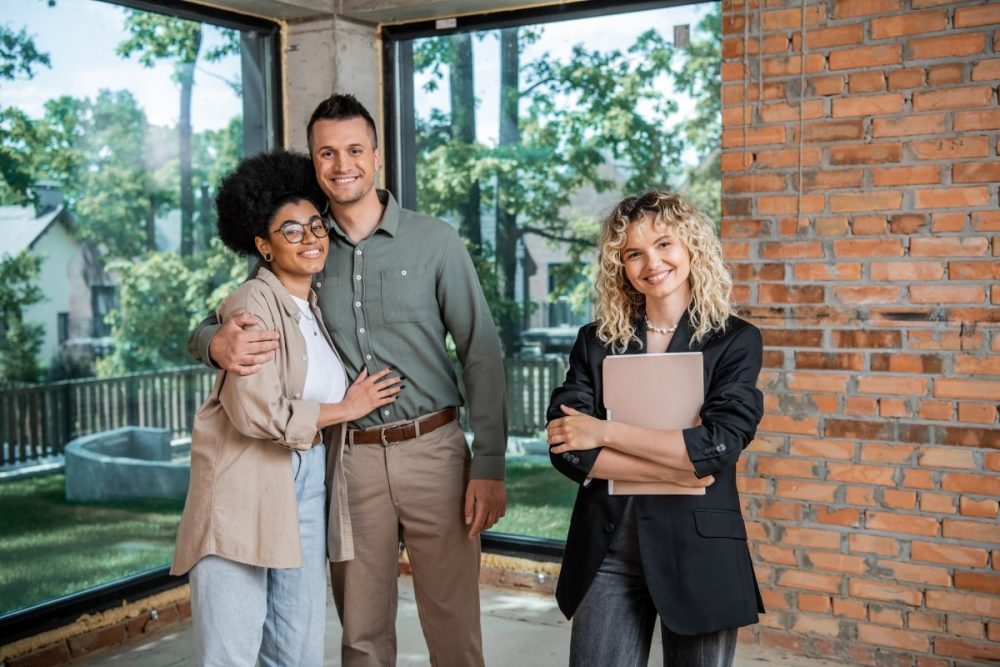 A smiling couple posing with a real estate agent in an unfinished home, symbolising the completion of a property purchase or successful home sale.