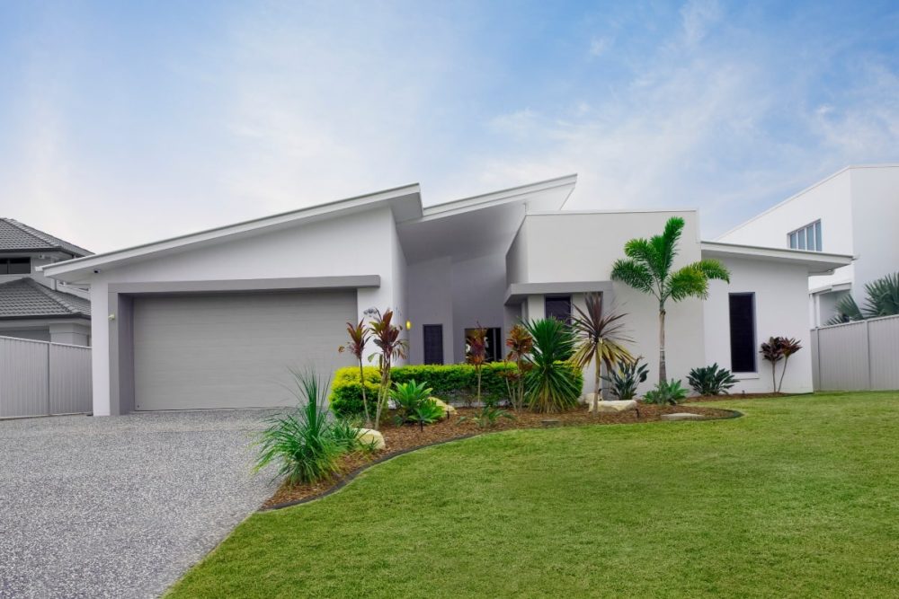 An immaculately staged house exterior with a modern design, showcasing a well-manicured lawn and a curated tropical garden. The inviting entrance and pristine white walls accentuate the property's appeal for sale.