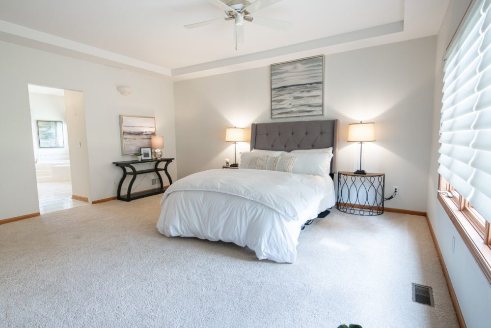 A master bedroom staged for sale, embodying a neutral colour palette that offers a calm and elegant atmosphere. Personal items have been removed to create a universal appeal, featuring a tasteful bed with white linen, complementary bedside tables with classic lamps, and a large window with blinds, all emphasizing the spacious and well-curated staging of the house for sale.