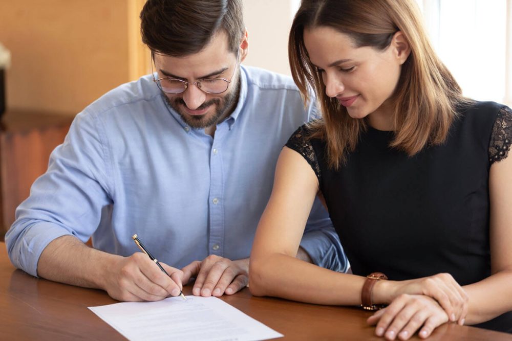 Close-up of a couple preparing to make an offer on a house, with the man signing a document and the woman overseeing the process.