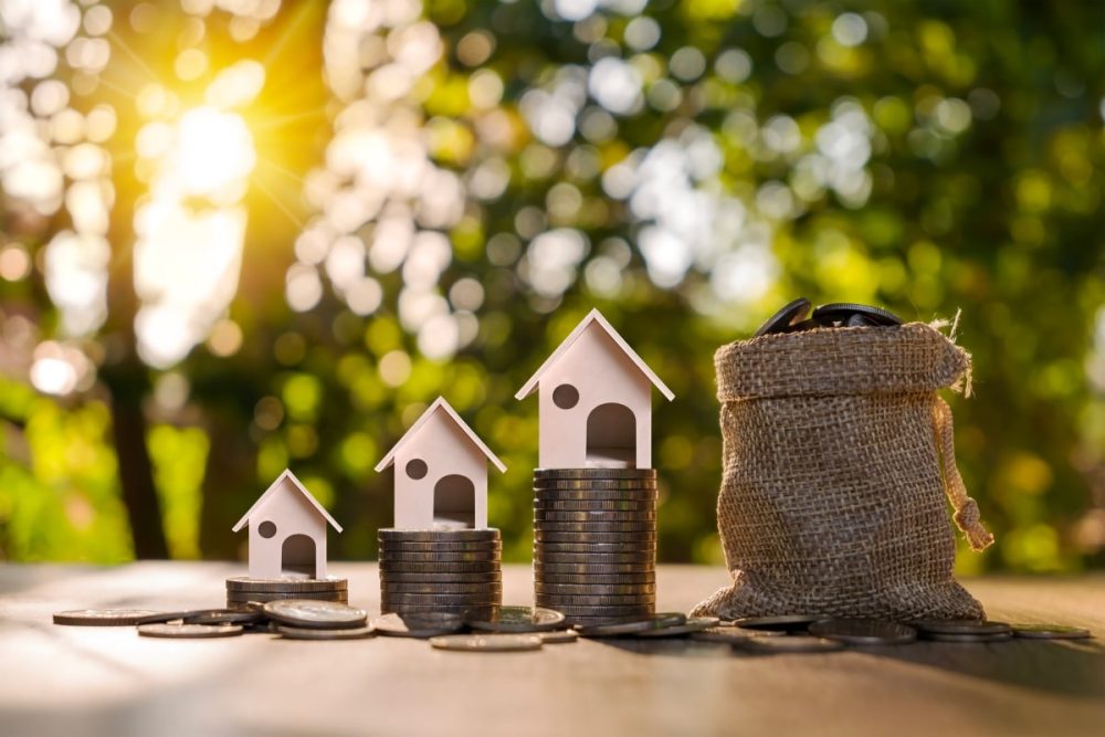 The progression of property investment shown with miniature houses placed on graduated coin stacks next to a money sack, representing the strategic financial steps in buying a second home.