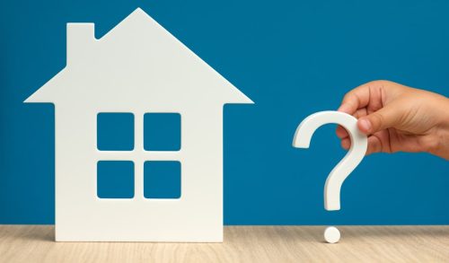A white cutout of a house on a blue background, with a hand holding a large white question mark next to it, symbolising the questions to ask when buying a house.