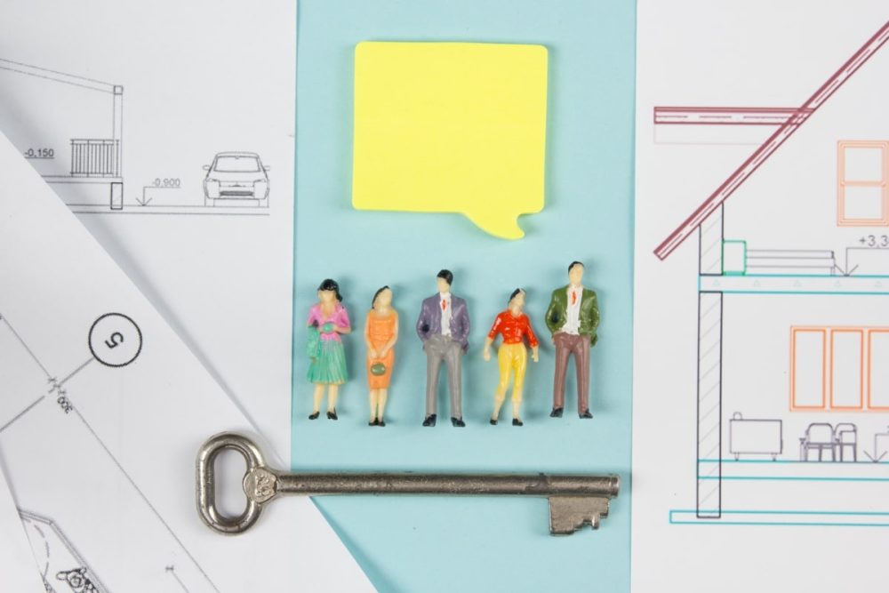 Miniature figurines lined up against a backdrop of house plans and a large key, representing the diversity of stakeholders and the complexity of questions to consider when buying a house.