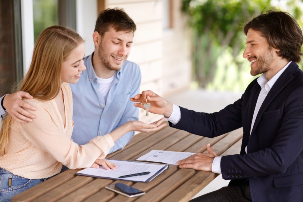 A professional real estate agent hands over keys to a happy couple, a moment that highlights the need to question the agent's success rate and experience.