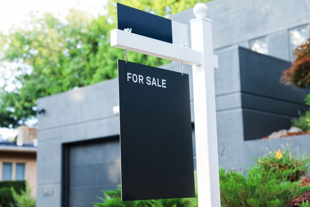 The image shows a stylish black and white 'For Sale' signpost, representing the initial step in marketing your property and prompting sellers to ask their real estate agent about listing visibility.