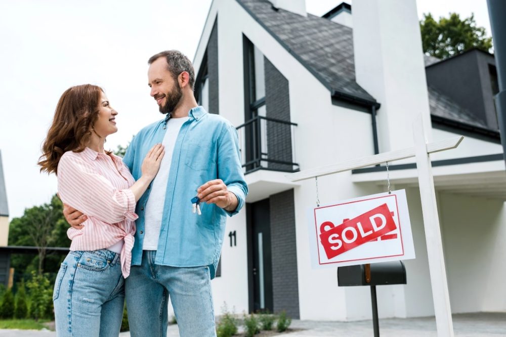 Happy couple celebrating their new home purchase, holding keys with a 'sold' sign in front of a modern house.