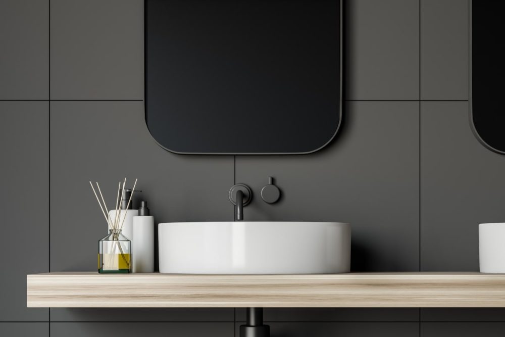 A sophisticated bathroom vanity staged for sale, featuring a sleek white basin atop a light wooden countertop against a dark tiled backdrop with a contemporary mirror. The scene is accented with a minimalist arrangement of bathroom accessories, including a diffuser and neatly placed toiletries, offering tips for staging for photos that enhance the appeal of the home.