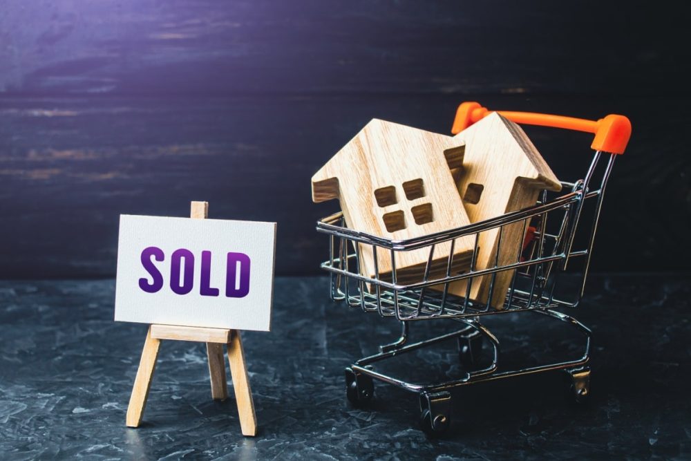 Miniature houses placed inside a small shopping cart with a 'SOLD' sign displayed, illustrating the scenario of buying a house before selling the previous home.